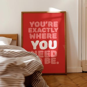 Può includere: Un poster rosso incorniciato con la scritta "YOU'RE EXACTLY WHERE YOU NEED BE." Il testo è in un carattere audace e giocoso, con la parola "YOU" in bianco. La cornice è marrone caldo e il poster è appoggiato a una parete bianca.