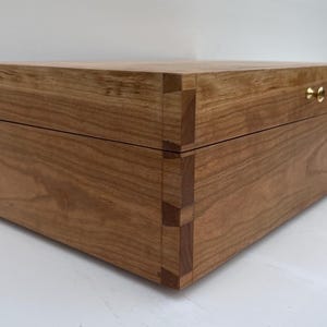 May include: A wooden box with a hinged lid and brass hardware. The box is made of light brown wood with a natural grain pattern.