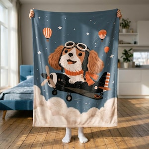 May include: A soft throw blanket featuring a brown and white spaniel dog wearing an aviator hat and orange scarf, piloting a black airplane through a dark blue sky with white stars, orange hot air balloons, and white clouds at the bottom.