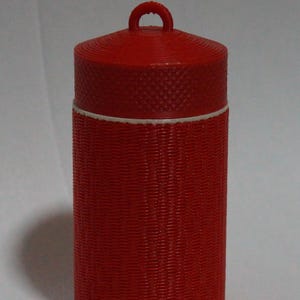 May include: A red cylindrical container with a textured, woven-like pattern. The container has a matching lid with a small handle. A white band separates the lid from the body of the container. The container is approximately 5 inches tall.
