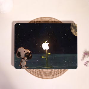 Könnte beinhalten: Ein Laptop-Aufkleber mit einer Grafik von Snoopy, der eine Kamera hält und ein leuchtendes, weißes Apple-Logo in Blumenform fotografiert. Die Szene zeigt einen dunklen, sternenklaren Nachthimmel und einen Teilmond über einem ruhigen Gewässer.