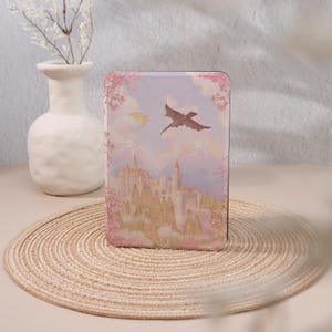 May include: A pink and purple illustrated book cover featuring a castle and two dragons flying in the sky. The castle is surrounded by trees and mountains.