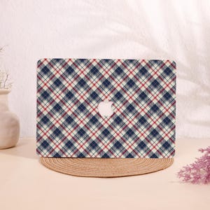 May include: A laptop case featuring a plaid pattern in navy blue, red, and cream. The case has a white Apple logo in the center. The laptop case is sitting on a round, woven mat.