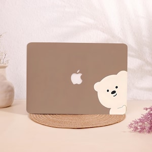 May include: A brown laptop with a white Apple logo and a white bear sticker peeking from the corner. The laptop is on a round, woven mat. The background is a light beige color.