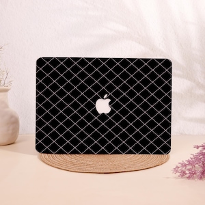 May include: Black laptop case with a quilted pattern and a white Apple logo. The case has a grid of white stitching over a black background. The laptop case is sitting on a round, woven mat.