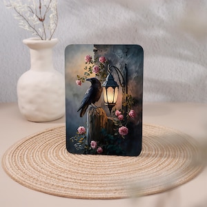 May include: A rectangular print featuring a black raven perched on a wooden post next to a glowing wall-mounted lantern. The scene is decorated with climbing vines and soft pink roses against a dark, textured background with muted blue tones.