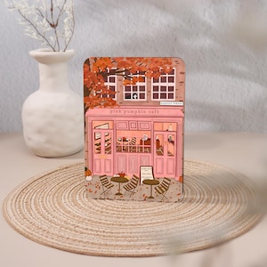 May include: A rectangular decorative sign featuring an illustration of a pink storefront labeled 'pink pumpkin café' and 'HARVEST STREET'. The scene includes autumn trees, a cat in a window, outdoor tables, chairs, and pumpkins on the ground.