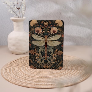 May include: A rectangular case with a dragonfly design. The dragonfly is set against a dark background with floral and leaf patterns in green, brown, and peach. The case is displayed on a woven mat.