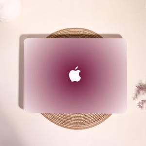 May include: A laptop with a smooth, matte finish featuring a gradient color scheme that transitions from a deep, dark magenta at the center to a lighter, pale pink at the edges. A white Apple logo is centered on the lid of the closed device.