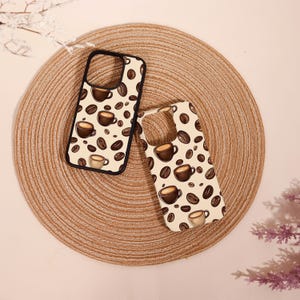 May include: Two phone cases with a coffee theme. The cases have a cream background with a pattern of coffee cups and coffee beans. One case has a black border. The cases are on a woven, circular mat.