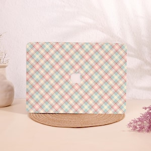 Könnte beinhalten: Eine Laptop-Hülle mit einem pastellfarbenen Karomuster in Rosa, Blau und Gelb. Die Hülle hat ein weißes Apple-Logo und liegt auf einem runden, gewebten Platzset. Der Hintergrund ist hellbeige.