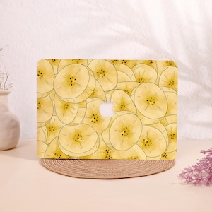 Könnte beinhalten: Eine Laptop-Hülle mit einem sich wiederholenden Muster aus gelben Bananenscheiben. Das Design beinhaltet das Apple-Logo in der Mitte. Die Hülle liegt auf einem runden, gewebten Platzset mit neutralem Hintergrund.