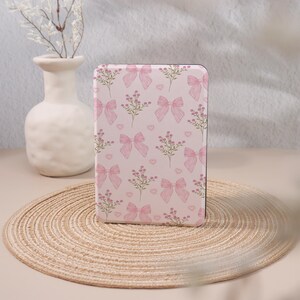 May include: A pink rectangular case adorned with a repeating pattern of pink bows, floral sprigs, and small hearts. The case is displayed against a neutral backdrop, with a vase of white flowers and a woven placemat.