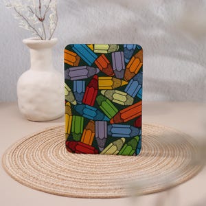 Peut inclure: Coque de téléphone rectangulaire avec un motif de crayons colorés sur fond vert foncé. Les crayons sont de différentes couleurs, dont le rouge, l'orange, le bleu et le jaune. L'étui est posé sur une surface beige.