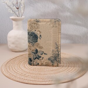 May include: A rectangular case featuring a vintage floral design with a large blue rose, butterflies, and botanical elements. The background has a faded, aged paper texture with printed text. The case is placed on a round, woven mat.