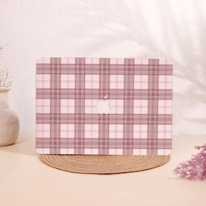 May include: A laptop with a pink and white plaid pattern. The laptop has a white Apple logo in the center. The laptop is on a round, woven placemat. The background is a light pink color.