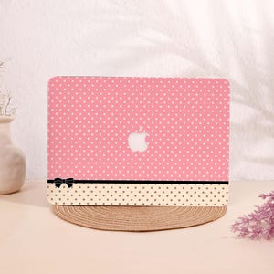 May include: Pink laptop case with white polka dots and an Apple logo. The case has a black ribbon and a beige dotted pattern at the bottom. The case is on a round, woven mat.