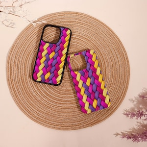 May include: Two phone cases with a colorful woven pattern in yellow, purple, and pink. One case is in a black frame. The cases are on a round, woven placemat. The background is a light beige.