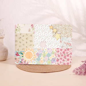 May include: A laptop case with a patchwork design featuring floral patterns, a watercolor sun, and gold star accents. The case has a cutout for the Apple logo and rests on a woven mat.