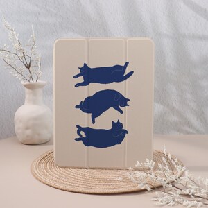 May include: A beige tablet case featuring three navy blue cat silhouettes in playful poses. The case is rectangular and has a folding cover. The design is simple and modern, suitable for cat lovers.