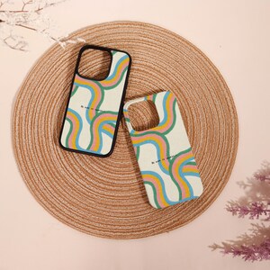 May include: Two phone cases with a colourful rainbow design and the text "Be kind to yourself." The cases have a cream background with a black border. The cases are on a woven, circular mat.