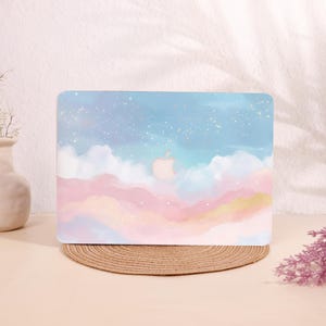 May include: A laptop case featuring a watercolor design of a dreamy sky with pastel pink, blue, and gold hues. The case has a light blue sky with white clouds and gold speckles. The Apple logo is visible in the center.