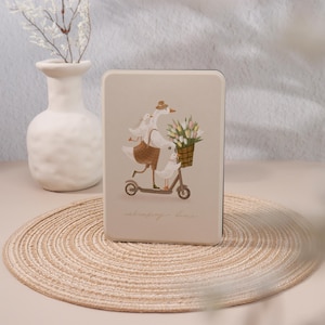 May include: A beige rectangular case featuring a whimsical illustration of geese riding a scooter, carrying flowers in a basket. The geese are dressed in hats and carrying bags. The case is on a woven mat.