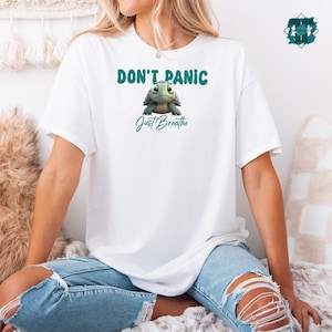 May include: White t-shirt with the text "DON'T PANIC" above a cartoon turtle and the words "Just Breathe" in teal. The turtle has a teal shell and a light green face. The shirt is worn by a person wearing ripped blue jeans.