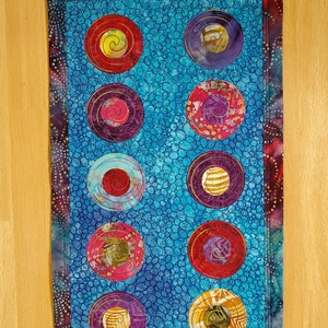 May include: A blue quilted wall hanging with a patterned fabric border. The center of the hanging features nine circular designs with swirling patterns in shades of red, purple, gold, and white.