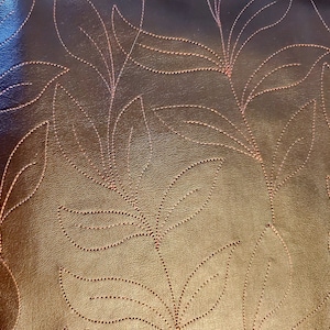 May include: Close-up of a dark brown leather surface with an embossed floral pattern. The design features stylized leaves and stems, created with dotted lines and a lighter brown color, creating a textured effect. The overall aesthetic is elegant and decorative.