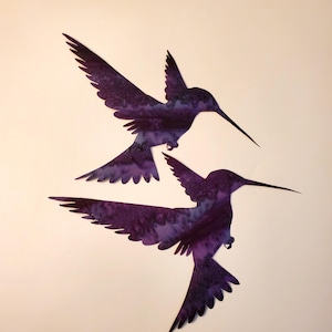 May include: Two flat, cutout hummingbird silhouettes in flight, featuring a deep purple and dark violet watercolor-style pattern with mottled textures across their wings and bodies, positioned diagonally against a plain light background.