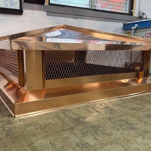 Op de afbeelding: Een koperen schoorsteenkap met een rechthoekig ontwerp. De kap heeft een gaasscherm aan de zijkanten en een schuine bovenkant. Het koper heeft een glanzende afwerking, die de omgeving weerspiegelt. De kap is ontworpen om bovenop een schoorsteen te zitten.