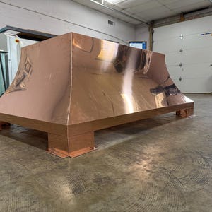 Puede incluir: Una gran campana extractora de color cobre con una base rectangular y una parte superior acampanada. La campana está hecha de metal brillante y se apoya sobre cuatro pies cuadrados. El diseño es moderno e industrial, adecuado para una cocina.