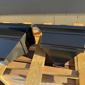 May include: Two black metal gutter downspout components, angled and trapezoidal in shape, are displayed on a wooden pallet. The components have a matte finish and are designed for efficient water drainage. The components are new.