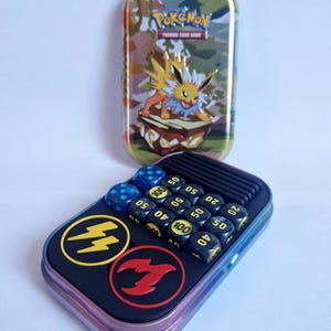 Könnte beinhalten: Eine rechteckige Pokémon Trading Card Game-Box mit Tarnmuster und einer gelb-orangenen Charakterillustration. Die Box ist geöffnet und zeigt Würfel und Spielsteine. Die Würfel sind schwarz mit weißen Zahlen.