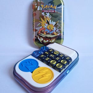 以下が含まれることがあります： カモフラージュ柄とサンダースのグラフィックが施された、ポケモンカードゲームのブリキ缶。 缶は開いており、サイコロ、青と黄色のトークン、白いトレイが表示されています。 サイコロは黒色で、数字は黄色です。