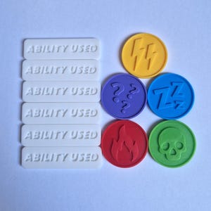 Puede incluir: Un conjunto de fichas y marcadores de juego. Marcadores rectangulares blancos con el texto "ABILITY USED". Fichas redondas: rayo amarillo, interrogaciones moradas, Z azules, llama roja y calavera verde.