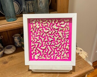May include: A white framed art piece featuring a vibrant pink background filled with numerous small, off-white, coral-like shapes. The artwork is displayed on a wooden surface, showcasing a unique, textured design.