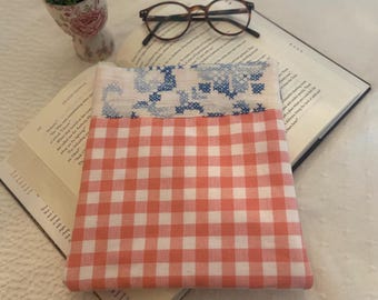 May include: A book cover made of a pink and white gingham fabric with a blue and white embroidered top section. The cover is lying on top of a book.