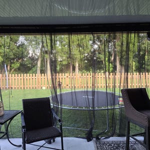 May include: A set of sheer black outdoor curtains hanging from a dark metal frame on a patio. The curtains are translucent, allowing a view of a backyard with a wooden fence, a trampoline, and outdoor furniture including chairs and a side table.