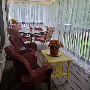 May include: A wicker patio set with red cushions and a yellow table. The set is on a wooden deck with white sheer curtains. There are potted plants with orange flowers on the table.