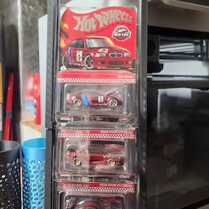 May include: A black display case holding five Hot Wheels die-cast model cars. The cars are various shades of red, with detailed designs and graphics. The top car has the Hot Wheels logo and "RED LINE" text. The case is made of black plastic.