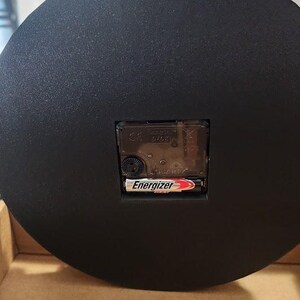 May include: Black round clock with a handle at the top. The back of the clock features a quartz movement and an Energizer battery. The clock is in a cardboard box.