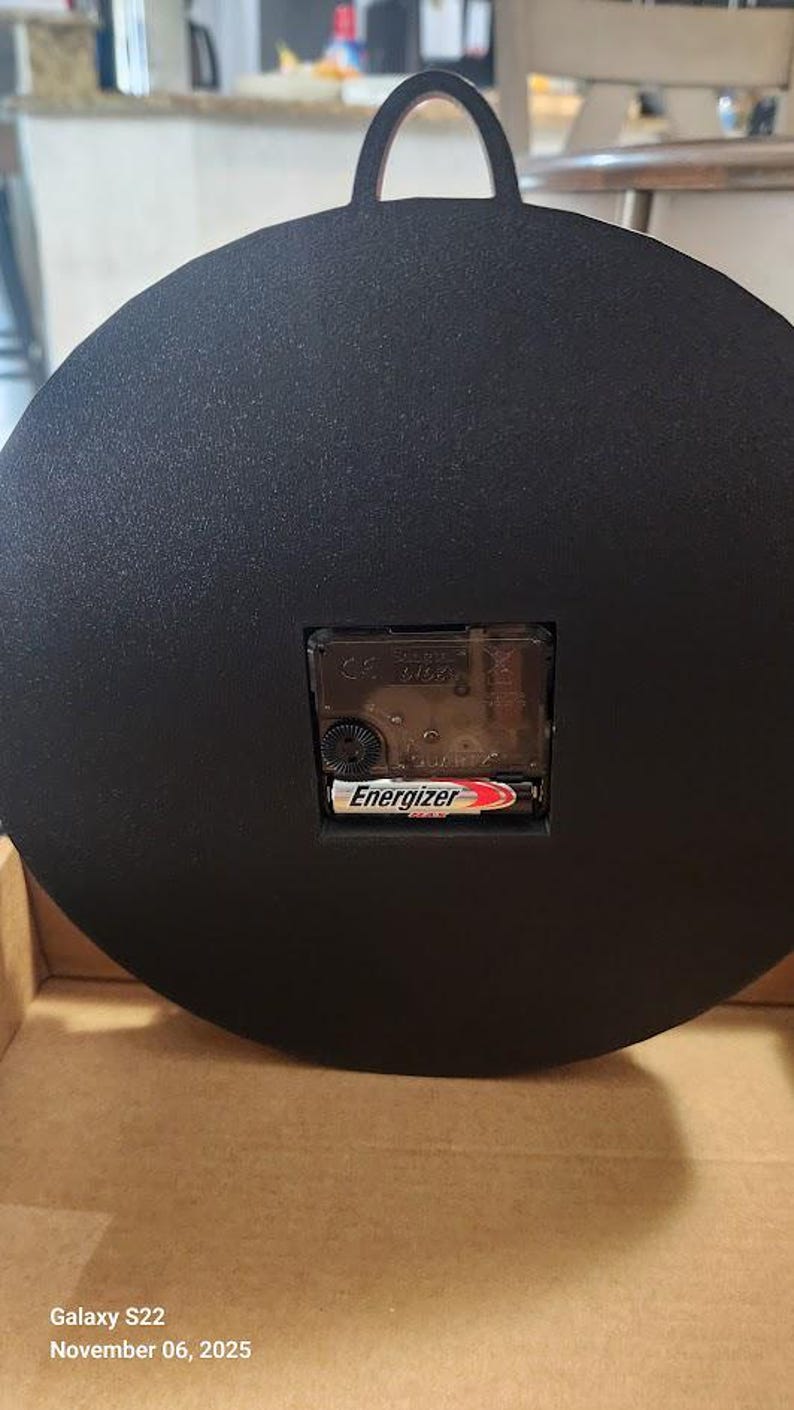 May include: Black round clock with a handle at the top. The back of the clock features a quartz movement and an Energizer battery. The clock is in a cardboard box.