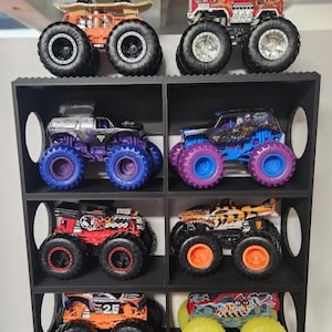 May include: A collection of toy monster trucks in various colors, including orange, red, blue, and silver, with large black tires. Some trucks have unique designs, such as a skateboarder on top or a tiger stripe pattern. The trucks are displayed in a black shelf unit.