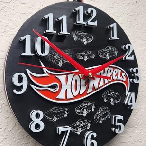 May include: Black Hot Wheels clock with white numbers and red hands. The clock face features Hot Wheels cars and the Hot Wheels logo in red and white. The clock is round and has a hanging loop.