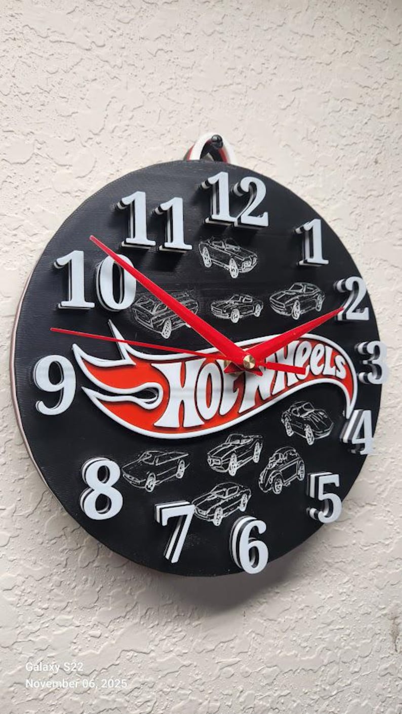 May include: Black Hot Wheels clock with white numbers and red hands. The clock face features Hot Wheels cars and the Hot Wheels logo in red and white. The clock is round and has a hanging loop.