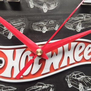 May include: A black, round clock with white numerals and red hands. The clock face features the Hot Wheels logo in white and orange, with car illustrations. The clock's hands are red, and the numbers are white.