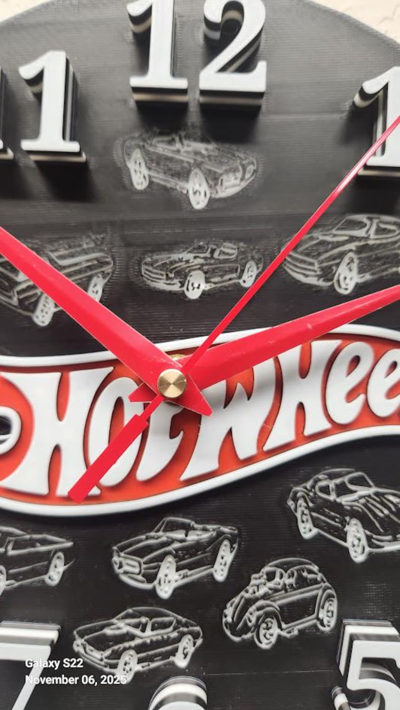May include: A black, round clock with white numerals and red hands. The clock face features the Hot Wheels logo in white and orange, with car illustrations. The clock's hands are red, and the numbers are white.