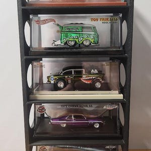 May include: A black display shelf holding five die-cast model cars in clear cases. The models include a gold M&M's Kool Kombi, a green Kool Kombi, a black and green truck, a purple 1972 Chevy Nova SS, and a blue 1959 Chevrolet Brookwood.